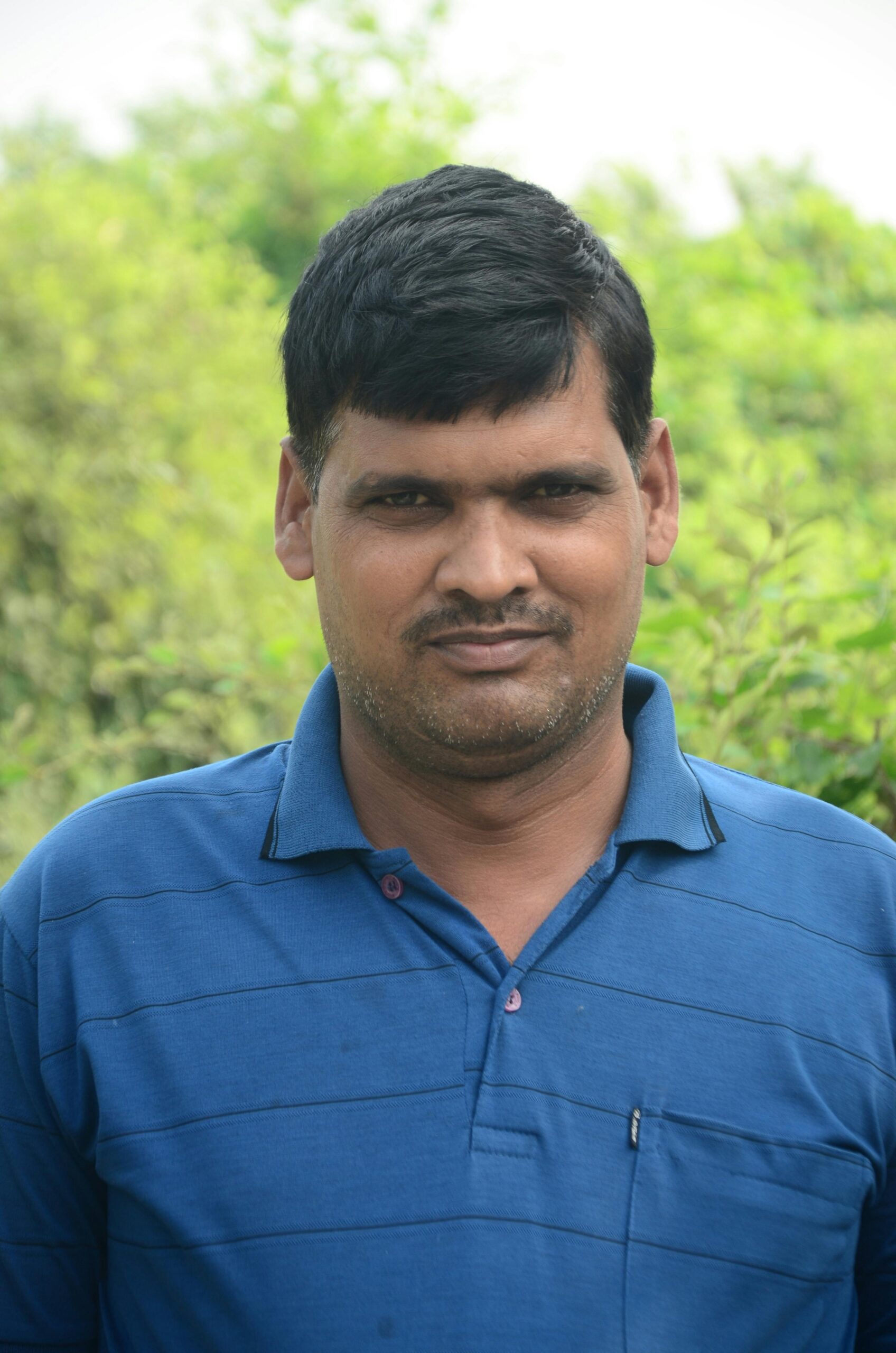 Casual portrait of an Indian man in a blue shirt with greenery in the background.